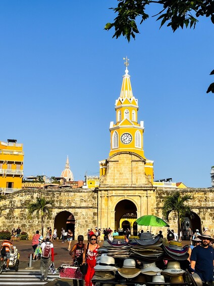 A historic yellow tower with a clock, surrounded by a bustling street with people and a vendor selling hats. 4