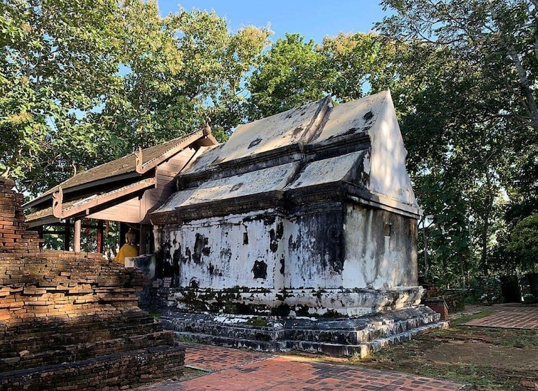 A weathered, white temple with a sloped roof, surrounded by trees and brick structures. 4