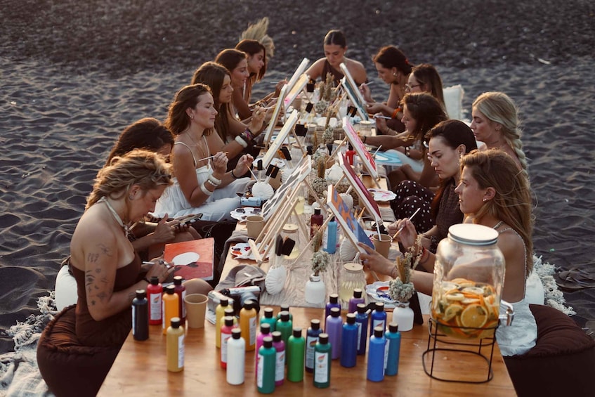 A group of people sitting around a table on a beach with various bottles and a jar of fruit. 4