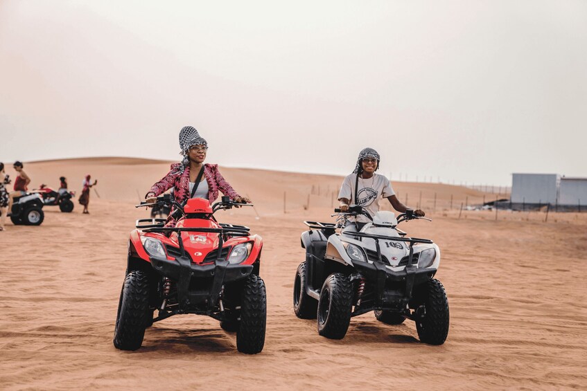 Two people riding ATVs in a desert with sand dunes and a fence in the background. 2