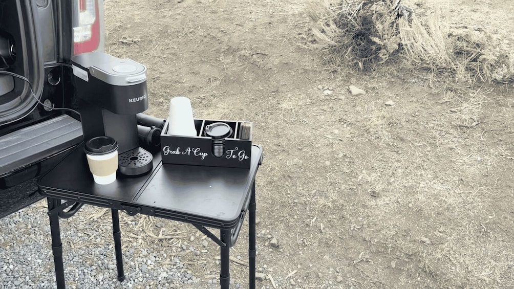 A portable coffee maker, a cup, and a tray with 'Grab A Cup To Go' on a small table. 3