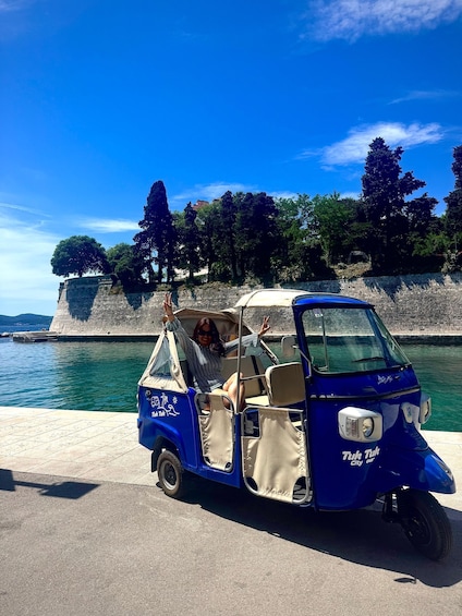 A blue tuk tuk parked by a waterfront with a stone wall and trees in the background. 5