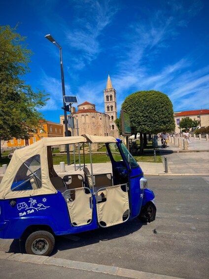 A tuk tuk parked on a street with a historic building and a tree in the background. 1