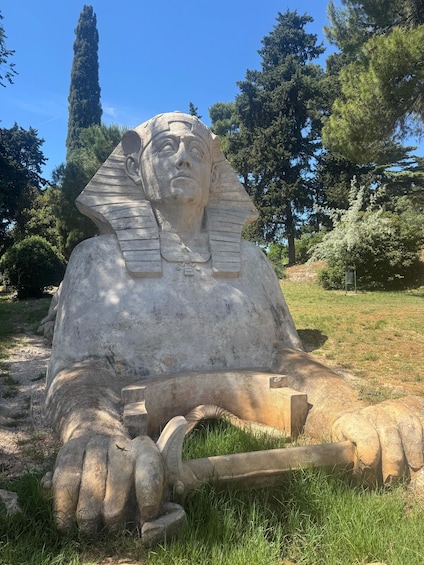 A large stone sphinx statue with a winged emblem, surrounded by grass and trees. 3
