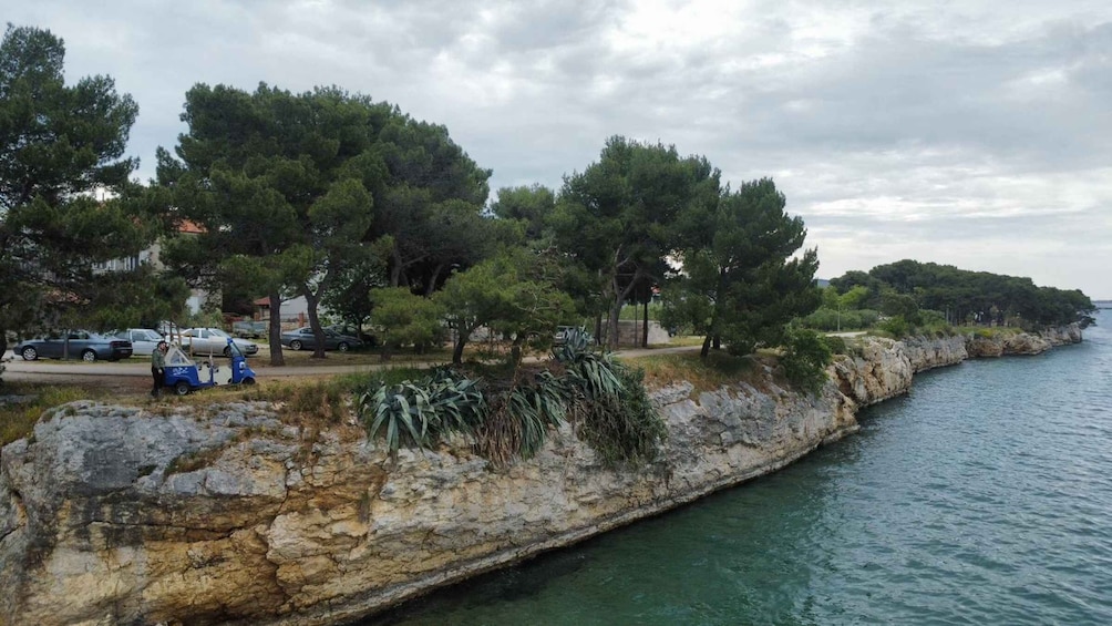 A coastal road with parked cars and a blue vehicle, trees, and a rocky shoreline. 4