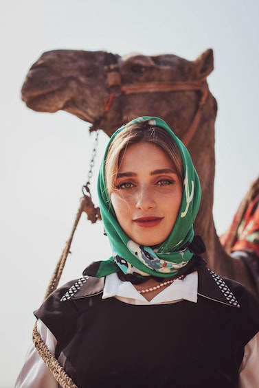 A person wearing a green headscarf and a black dress with a white collar, standing in front of a camel. 3