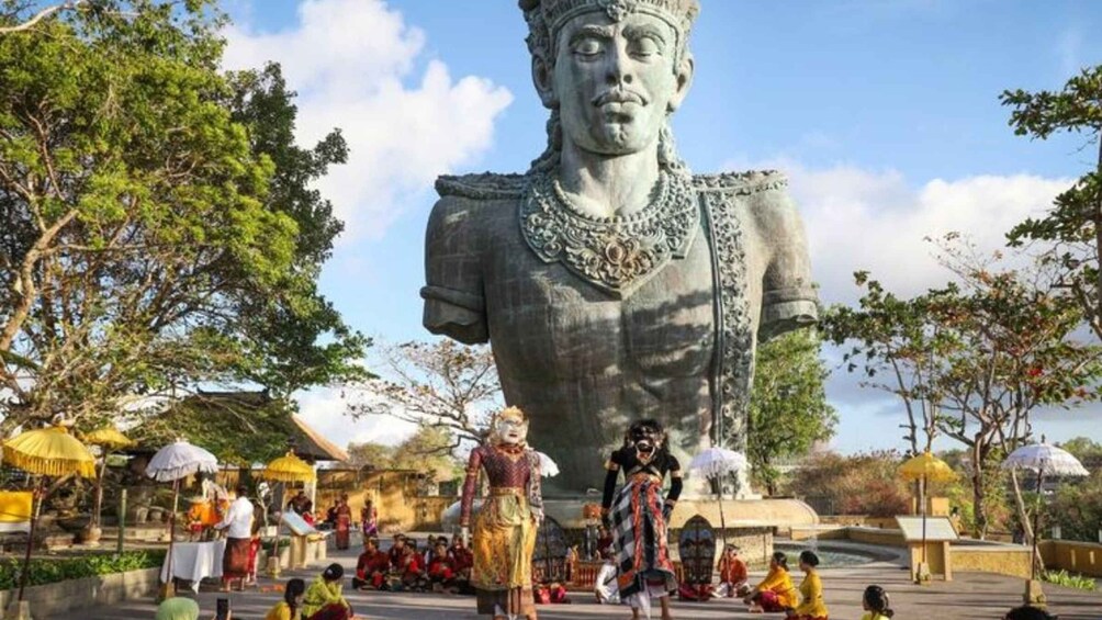 A large statue of a historical figure, surrounded by people in traditional attire, in a park-like setting with trees and umbrellas. 3