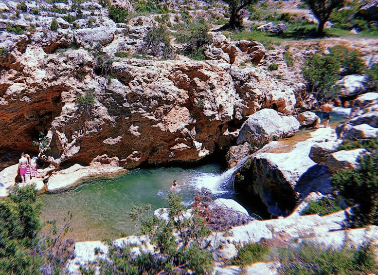 A rocky canyon with a clear stream, people swimming, and sparse vegetation. 3