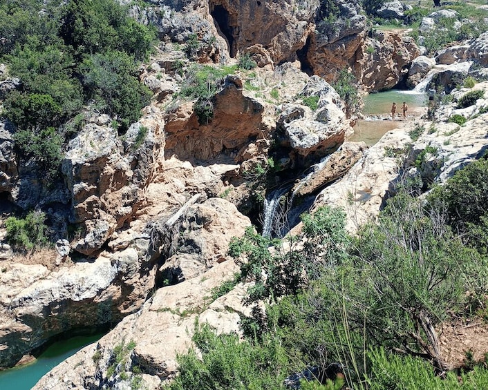 A rocky canyon with a waterfall and a small group of people near the water. 1