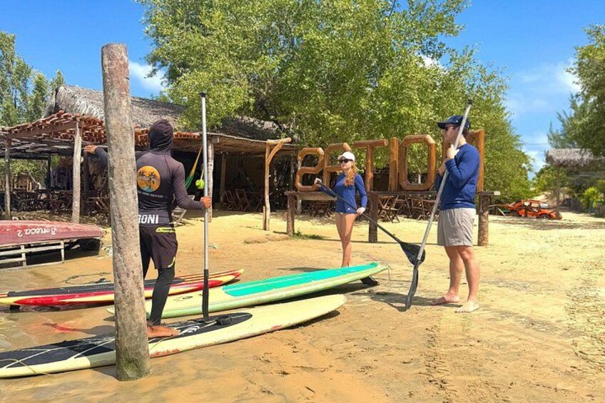 People on a sandy beach with surfboards and paddleboards, a wooden structure, and trees in the background. 4
