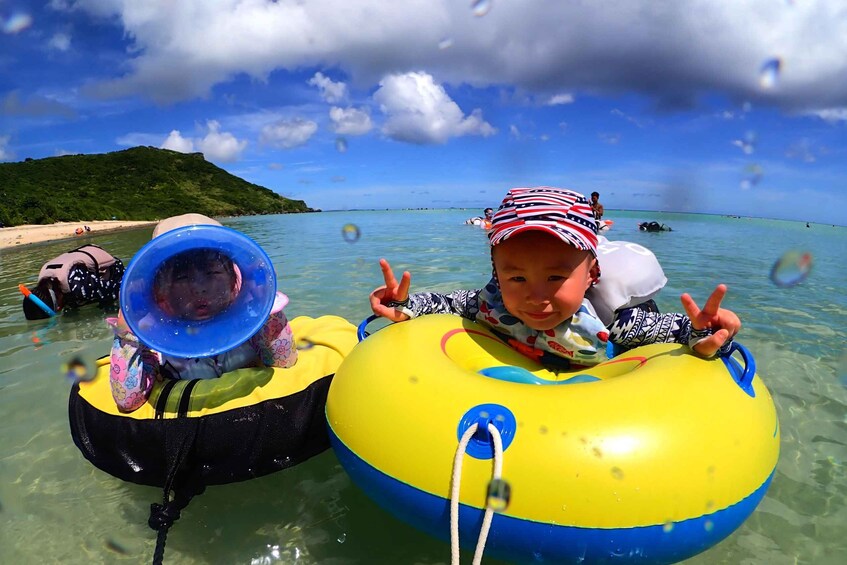 Two people floating on inflatable rings in clear water with a beach and hills in the background. 2