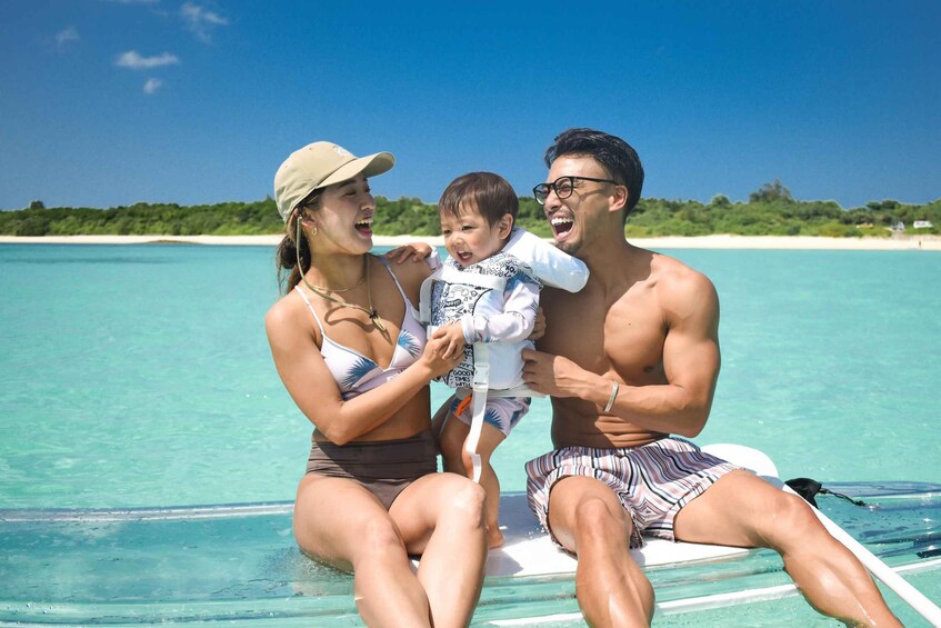 A family on a boat in clear turquoise water with a beach and greenery in the background. 3