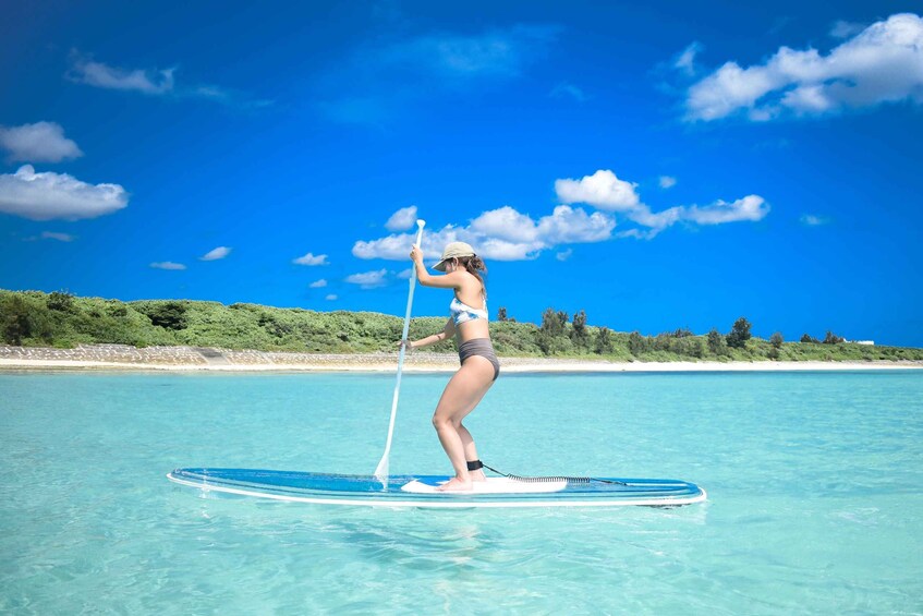 A person paddleboarding on clear turquoise water with a beach and greenery in the background. 5