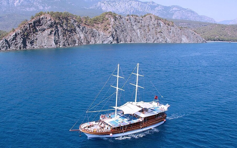 A large sailing boat with multiple decks and a flag, sailing on a calm sea with rocky cliffs in the background. 5