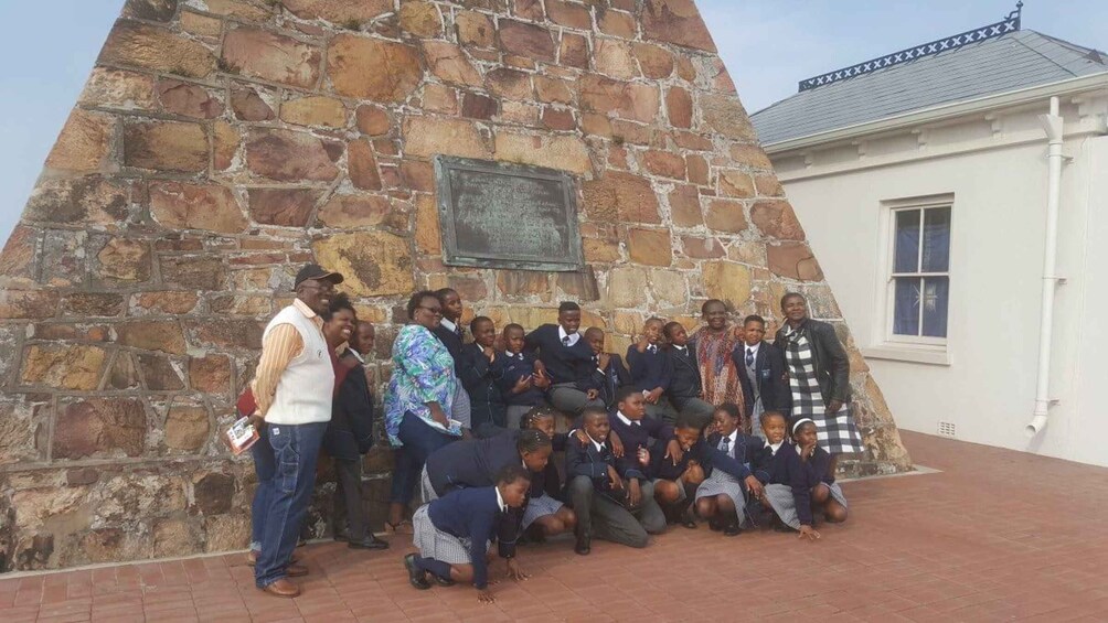 A group of people gathered around a stone monument with a plaque, in front of a building with a white facade and a grey roof. 1