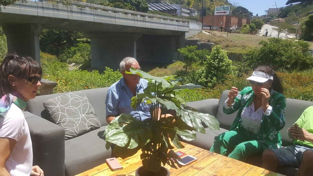 A group of people sitting on a couch outdoors with a large plant in the foreground and a bridge in the background. 3