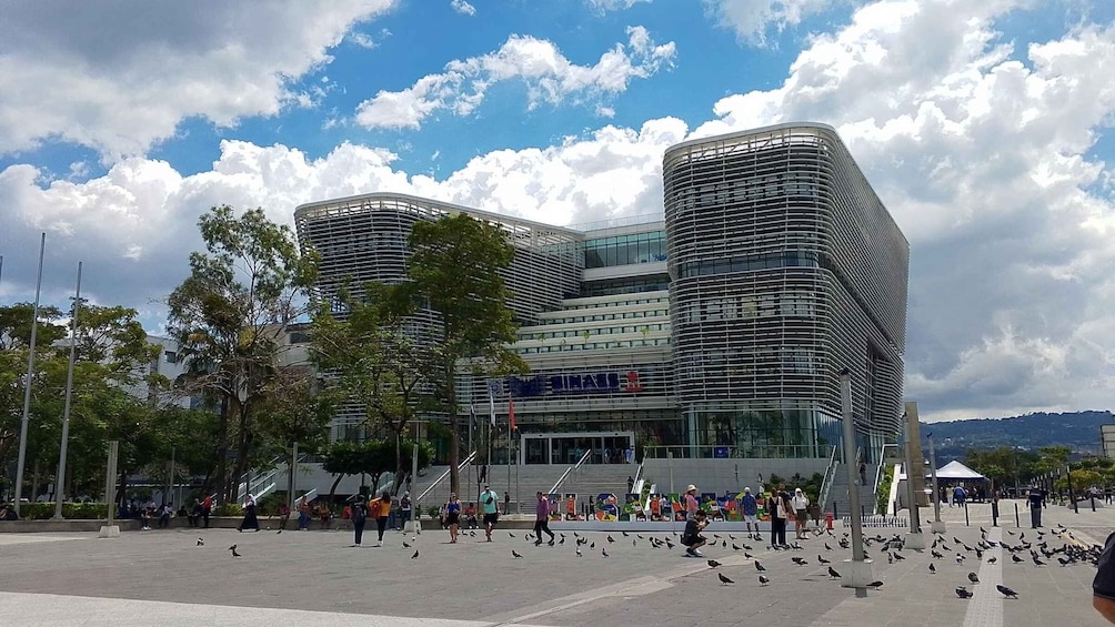 A modern building with a curved facade and a large glass entrance, surrounded by a plaza with people and pigeons. 1
