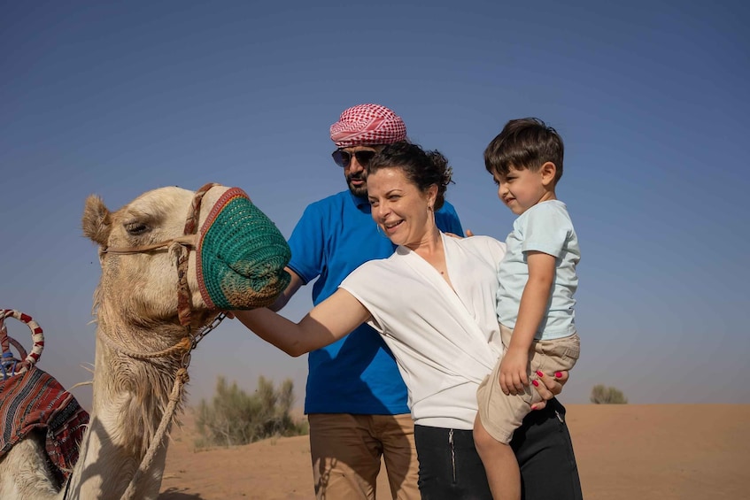 A camel with a saddle, a person in a blue shirt, and a child in a white shirt. 1