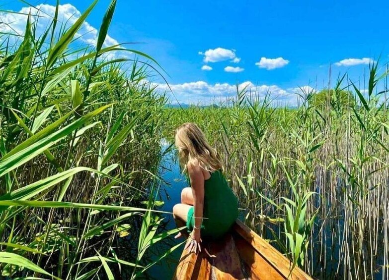 A person sitting on a wooden boat in a marshy area with tall grasses and a clear blue sky. 1