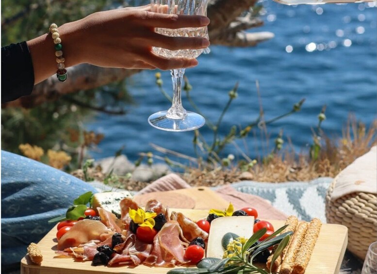 A person holding a wine glass, a charcuterie board with various meats and cheeses, and a body of water in the background. 5