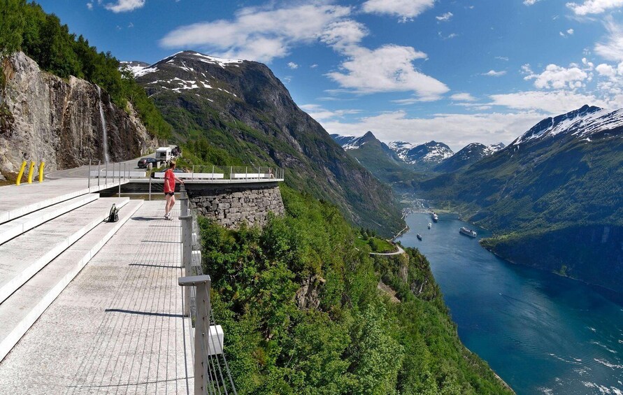 A scenic overlook with a person standing on a viewing platform, overlooking a fjord with mountains and a clear sky. 3
