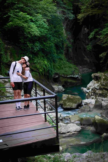 A couple standing on a wooden bridge over a river with rocks and greenery. 1