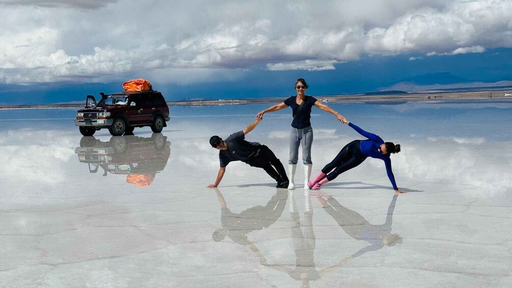 Three people performing a handstand on a reflective salt flat with a vehicle in the background. 4
