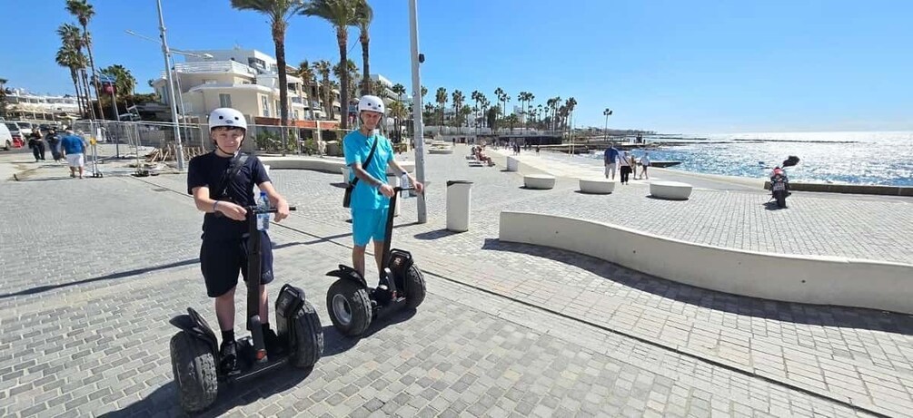 Two people on segways, a paved promenade, palm trees, and a beach in the background. 2