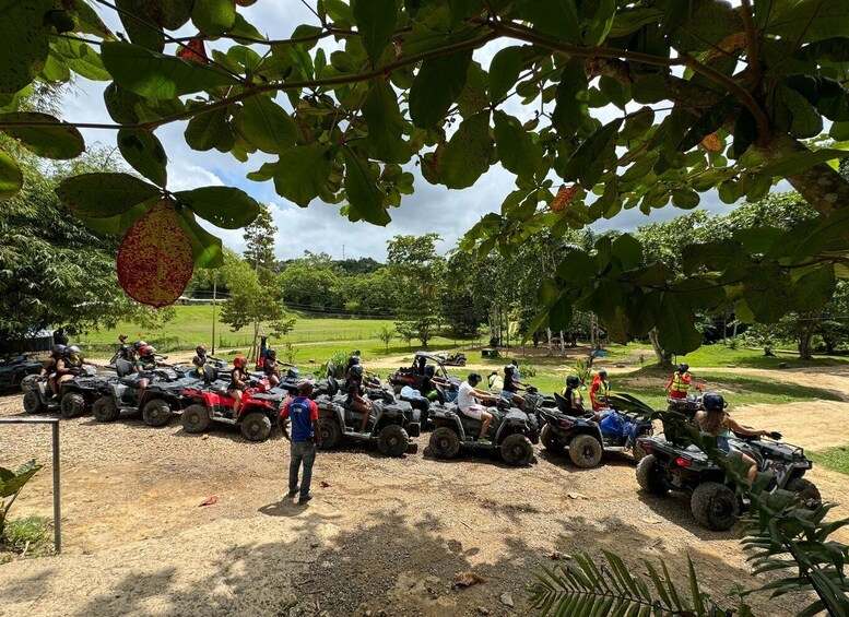 A group of people on ATVs lined up on a dirt path in a green, open area with trees and a clear sky. 3