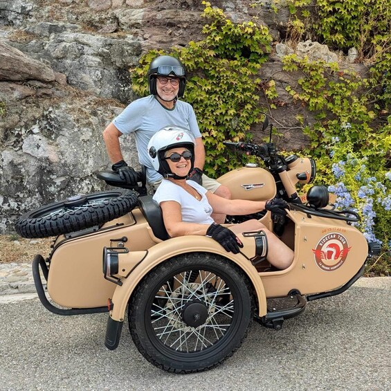 A vintage sidecar with two people, one driving and one sitting, parked on a paved area with greenery and rocks in the background. 3