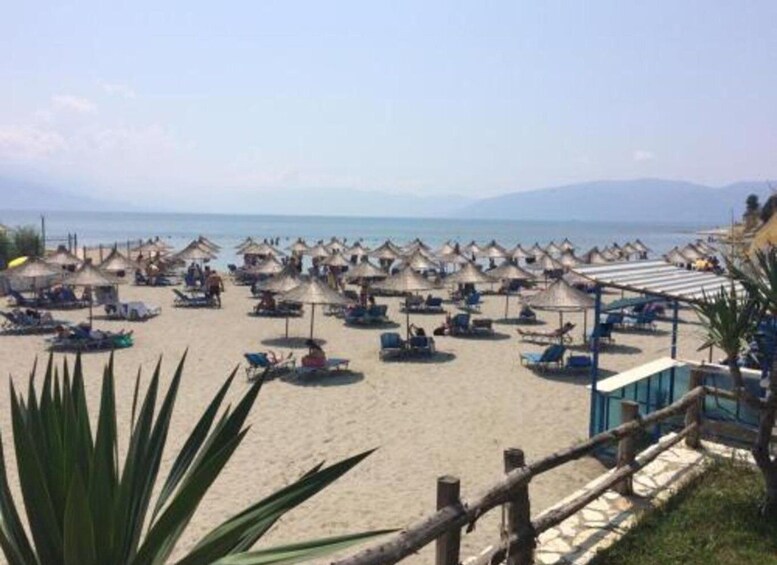 A beach with numerous sun umbrellas and lounge chairs, a wooden fence, and a clear view of the sea and distant mountains. 2
