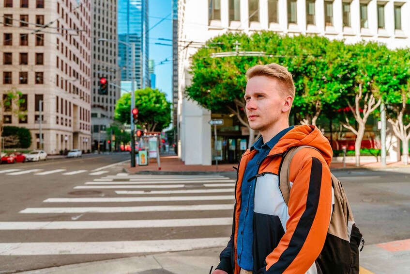A person standing at a crosswalk in an urban setting with buildings and trees in the background. 3
