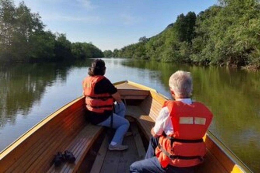 Two people in a narrow wooden boat wearing life jackets on a calm river surrounded by dense greenery. 1