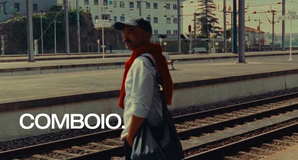 A man standing on a train platform with tracks, a cityscape in the background, and the word 'COMBOIO' visible. 2