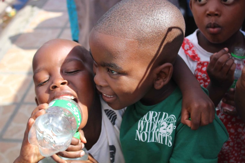 A child drinking from a bottle, another child wearing a green shirt with a logo, and a third child in a floral dress. 2