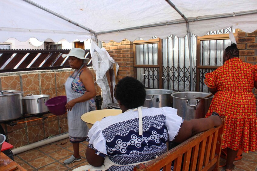 A group of people in a covered outdoor area with cooking pots and a brick wall. 5