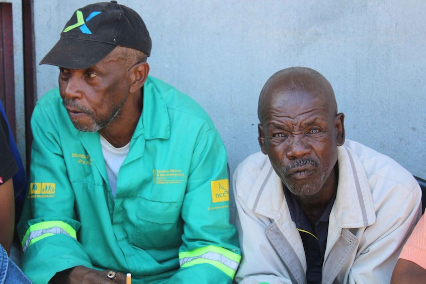 Two men sitting, one in a green jacket with a cap, and the other in a light-colored jacket. 3