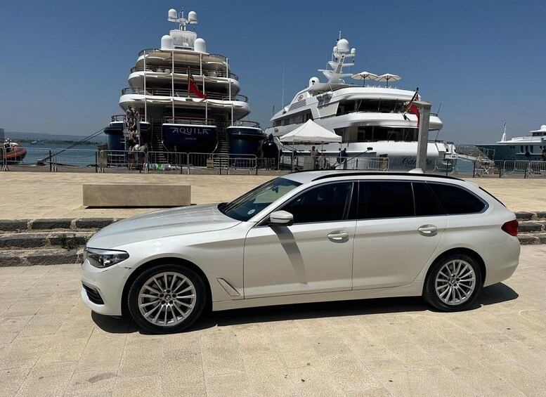 A white station wagon parked in front of a marina with yachts. 5