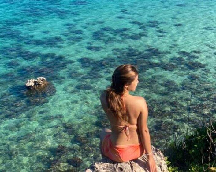 A person sitting on a rock by clear blue water. 1