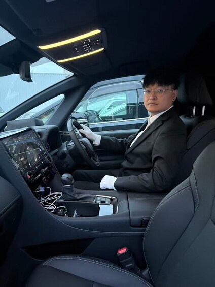 A person sitting in a car with a steering wheel, dashboard, and gear shift. 1