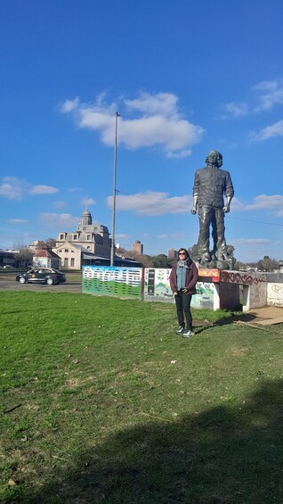 A person standing in front of a statue on a grassy field with buildings and a clear sky in the background. 5