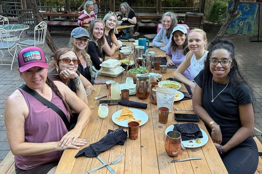 A group of people sitting around a wooden table outdoors, enjoying a meal with various dishes and drinks. 2