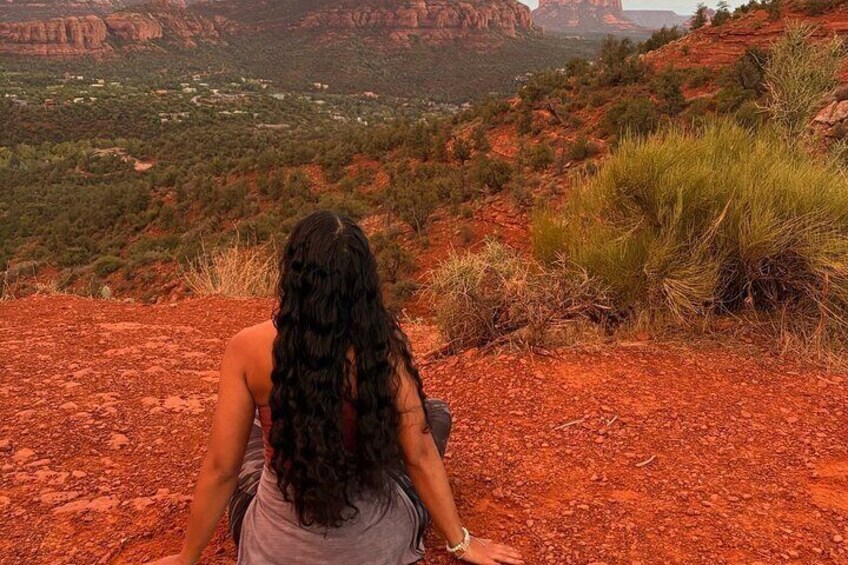 A person sitting on a red rock outcrop overlooking a valley with sparse vegetation and distant buildings. 5