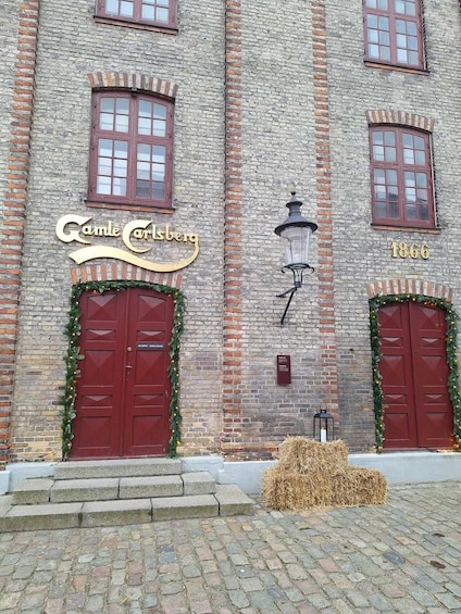 A brick building with a sign reading 'Gande Carlsberg' and the year '1866' above two red doors, with a lantern and hay bales in front. 2