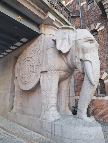 A stone elephant sculpture with a decorative emblem on its side, set against a brick building with a metal balcony. 5