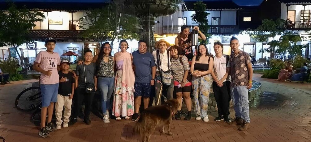 A group of people standing on a paved area with a dog in the foreground, trees, and buildings in the background. 5