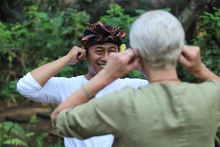 A person in a white shirt with a flower in their hair, standing in a forested area. 1