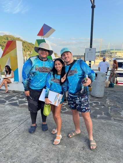 Three people wearing matching blue shirts with 'Riviera Charter Panama' printed on them, standing on a concrete surface with a marina and boats in the background. 5