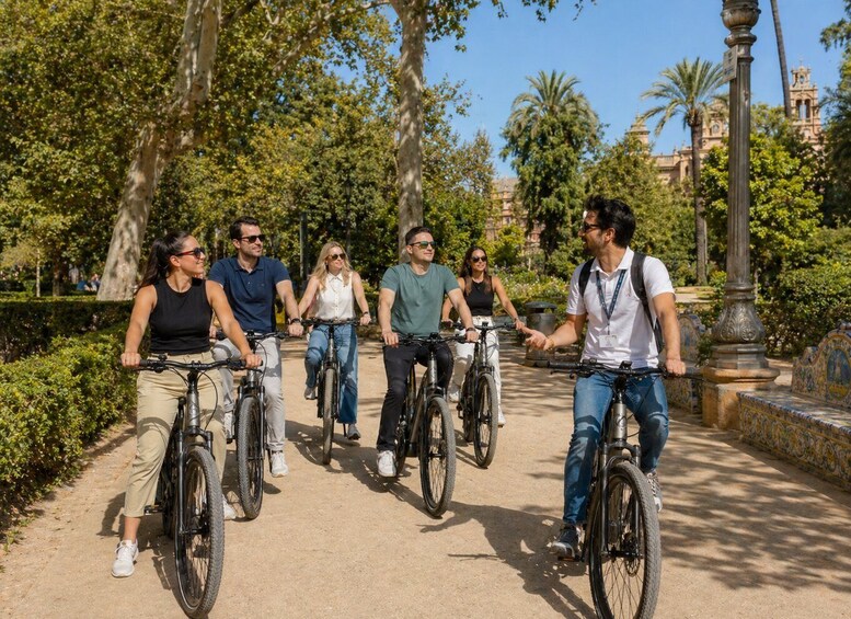 A group of people riding bicycles on a paved path in a park with trees and a building in the background. 2
