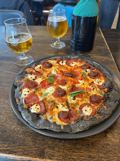 A pizza with pepperoni and cheese on a black plate, accompanied by two glasses of beer and a tumbler on a wooden table. 5